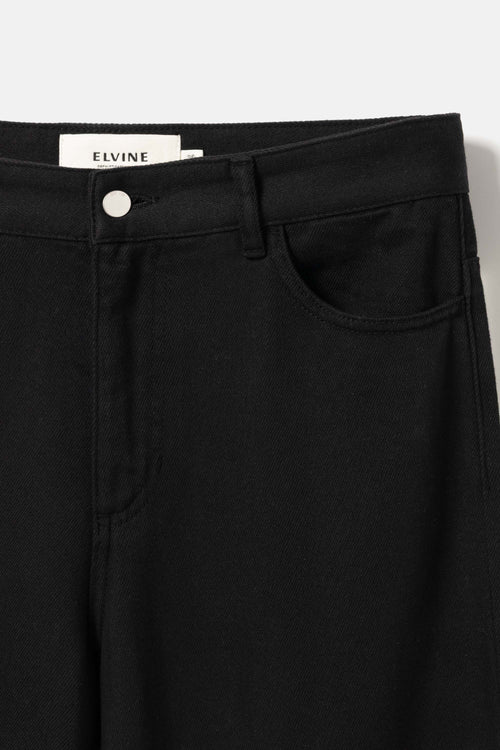 Close-up of black denim pants showing the waistband, button closure, belt loops, front pocket, and a visible ELVINE brand label.