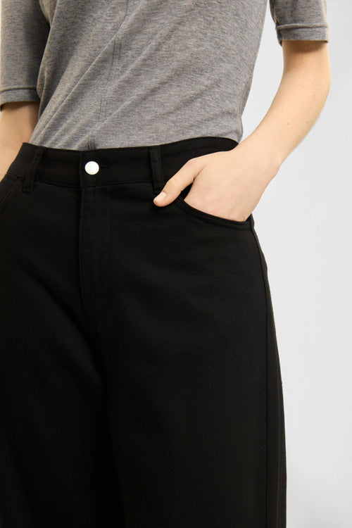 Close-up of a person wearing black wide-leg trousers and a heather gray short-sleeve top, with one hand in the trouser pocket, against a light gray studio background.
